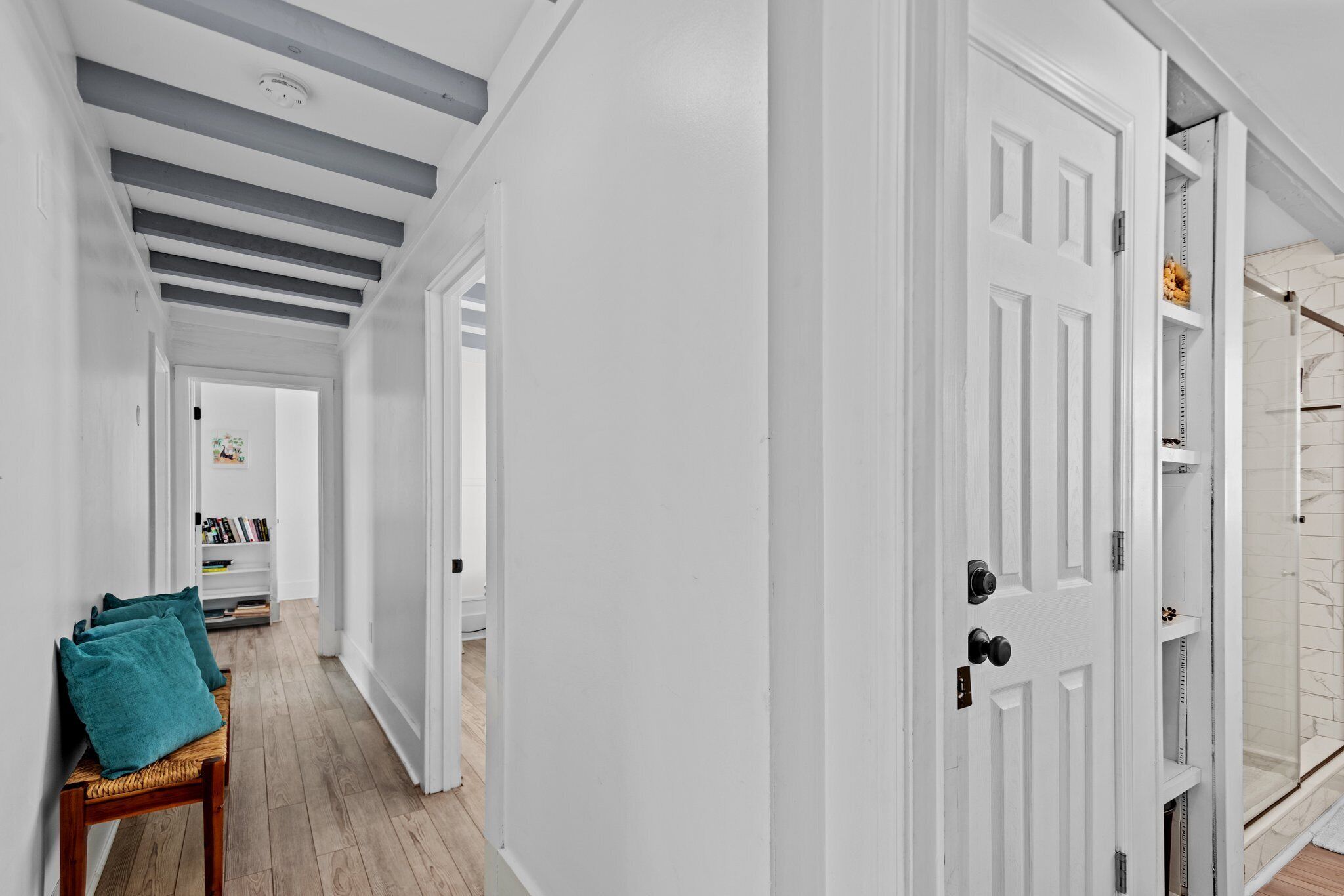 A hallway with white walls, a wooden bench with teal cushions, and a white door leading to a bathroom with a glass shower.
