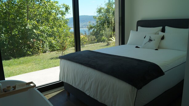 Un dormitorio con una cama grande, ropa de cama blanca y vistas a un jardín exuberante y un paisaje lejano a través de amplios ventanales.