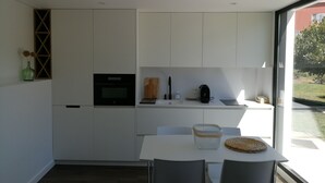 Una cocina compacta con armarios blancos, horno integrado, cafetera y un comedor con mesa y sillas.
