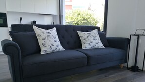 Un sofá gris oscuro con dos cojines estampados, ubicado en una habitación moderna con suelo de madera y un ventanal que ofrece vistas al exterior con vegetación.
