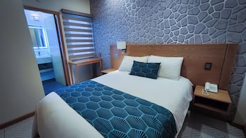 A hotel room with a large bed, a wooden headboard, a patterned wall, a bedside table with a phone, and a door leading to a bathroom.