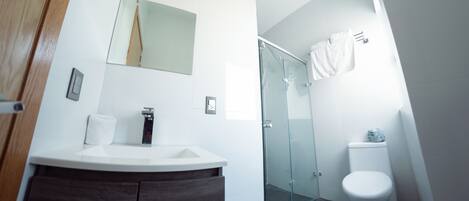 Un baño moderno con lavamanos blanco, tocador de madera oscura, cabina de ducha con mampara de vidrio y inodoro blanco.