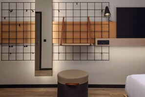 A modern hotel room with a wooden floor, a bed, a small round ottoman, a wall-mounted TV, and a grid-patterned mirror. - Moxy Lisboa Oriente (Lisbon)