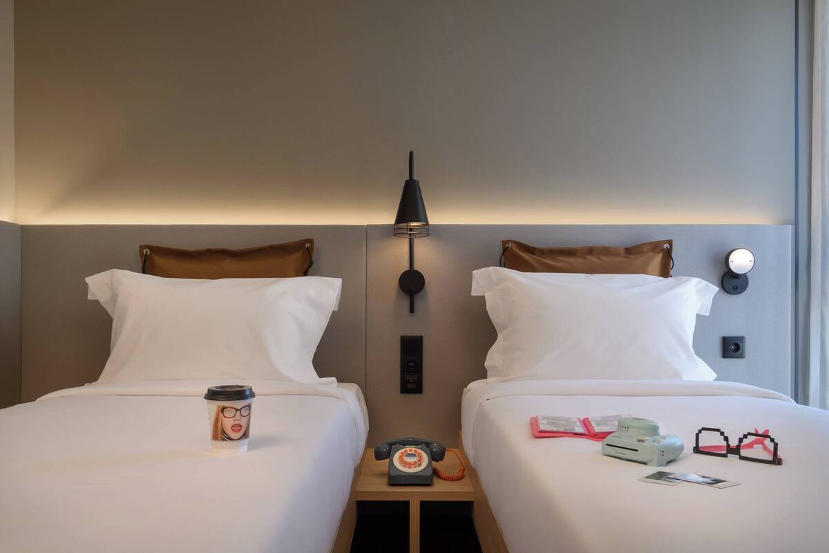 A hotel room with two beds, a bedside lamp, a telephone, a coffee cup, and a pair of glasses on the bed.