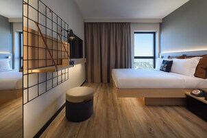 Quarto de hotel moderno com cama, pequena escrivaninha, TV fixada na parede, janela com cortinas e vista para a cidade.