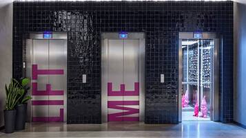 Three elevators with digital displays, a tiled wall with a geometric pattern, and a hallway with a potted plant.