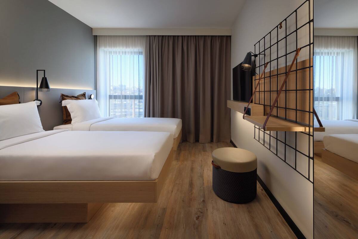 A modern hotel room with two beds, a large window with curtains, a wooden floor, and a wall-mounted shelf with a lamp.