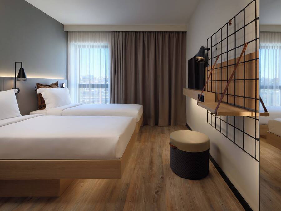 A modern hotel room with two beds, a large window with curtains, a wooden floor, and a wall-mounted shelf with a lamp.