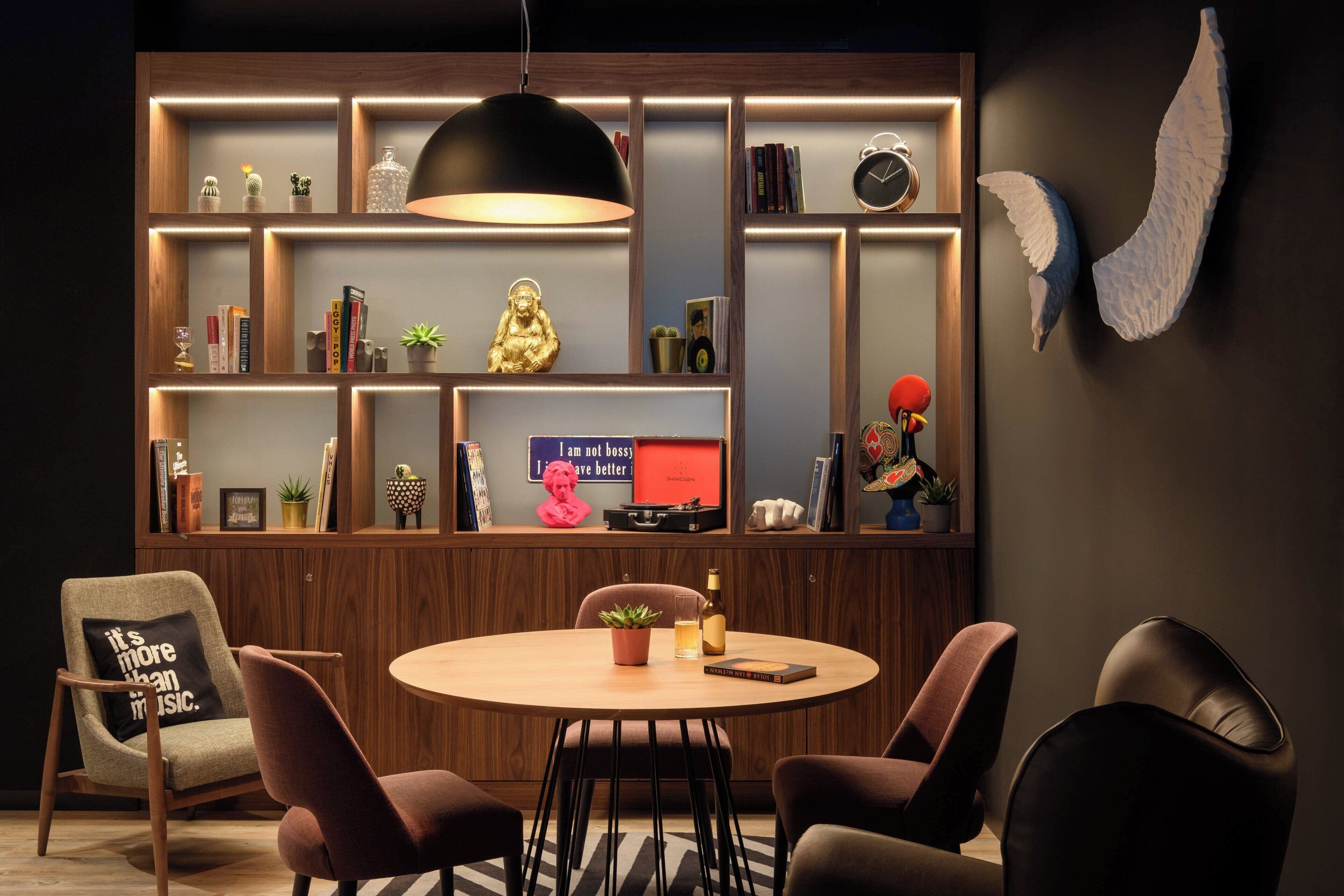 Una sala de estar moderna con una mesa redonda de madera, cuatro sillas, una estantería con diversos objetos decorativos y un aplique de pared.