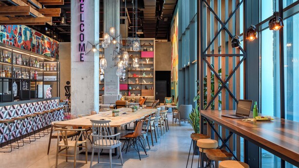 A modern bar area with a 'WELCOME' sign, a long wooden table with chairs, and a bar stocked with bottles. - Moxy Lisboa Oriente (Lisbon)