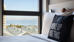 A hotel room with a bed, a pillow with text, a window with a view of buildings and water. - Moxy Lisboa Oriente (Lisbon)