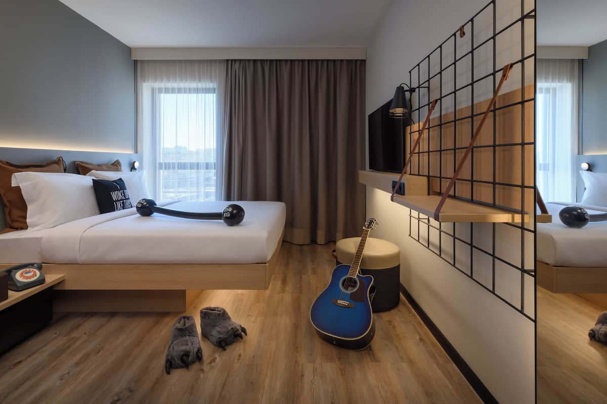 A modern hotel room with a bed, a guitar, a small stool, and a TV mounted on a grid wall.