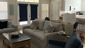 A spacious living room with a high ceiling, a ceiling fan, and a view of the kitchen through an open door.