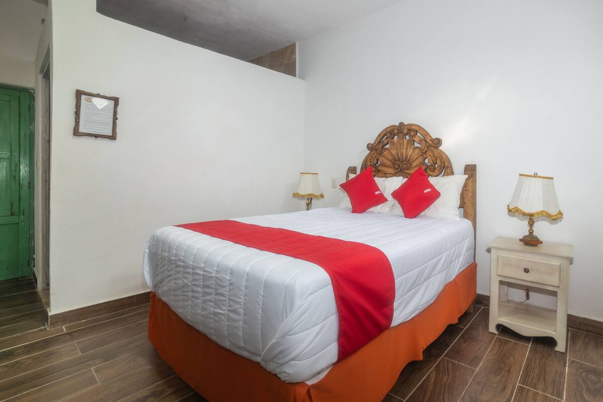 Een net opgemaakt bed met een wit en rode sprei, twee rode kussens en een houten hoofdbord. Er staat een nachtkastje met een lamp bij, en aan de muur hangt een lijst met een certificaat.
