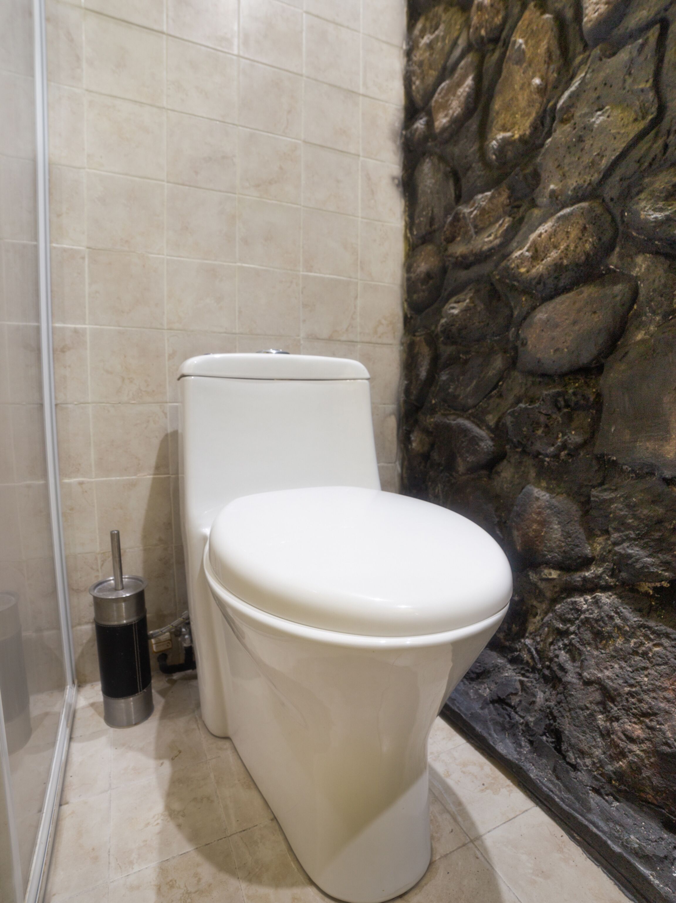 A bathroom with a white toilet, a black trash bin, and a stone wall.