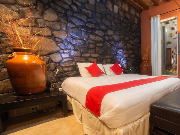 A bedroom with a stone wall, a large vase, and a bed with red pillows.