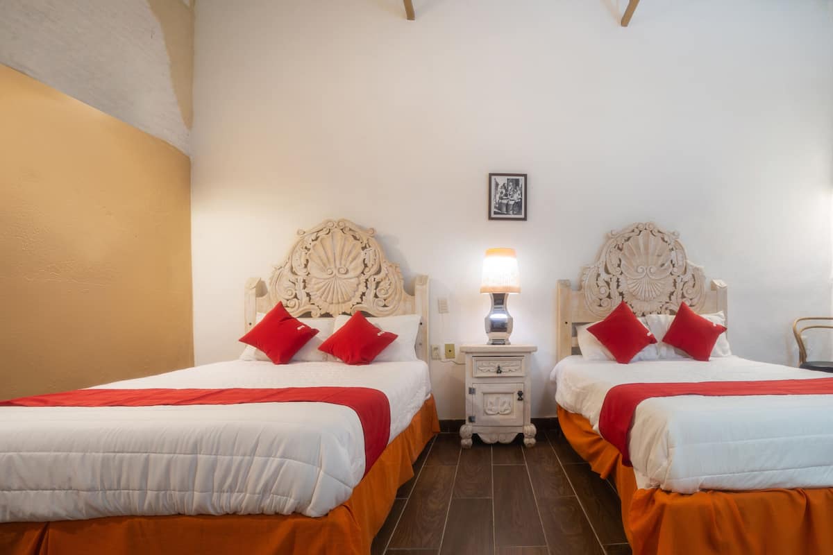 Een hotelkamer met twee bedden, elk voorzien van een sierlijk hoofdbord, witte beddengoed en rode kussens. Daartussen staat een nachtkastje met een lamp.