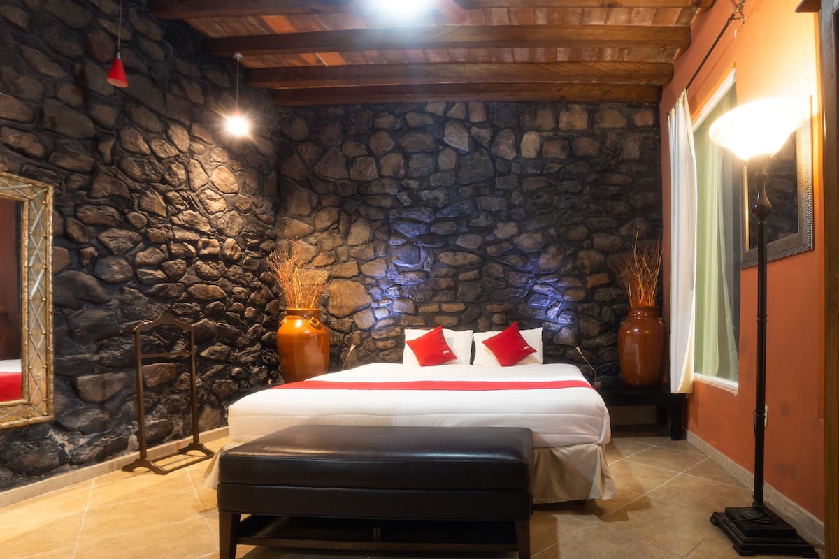 Een slaapkamer met een stenen muur, een bed met witte beddengoed en rode kussens, een bankje en een spiegel aan de muur.