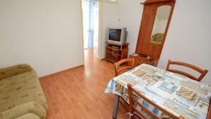A small room with a wooden floor, a green sofa, a dining table with chairs, a television on a stand, and a mirror on the wall.