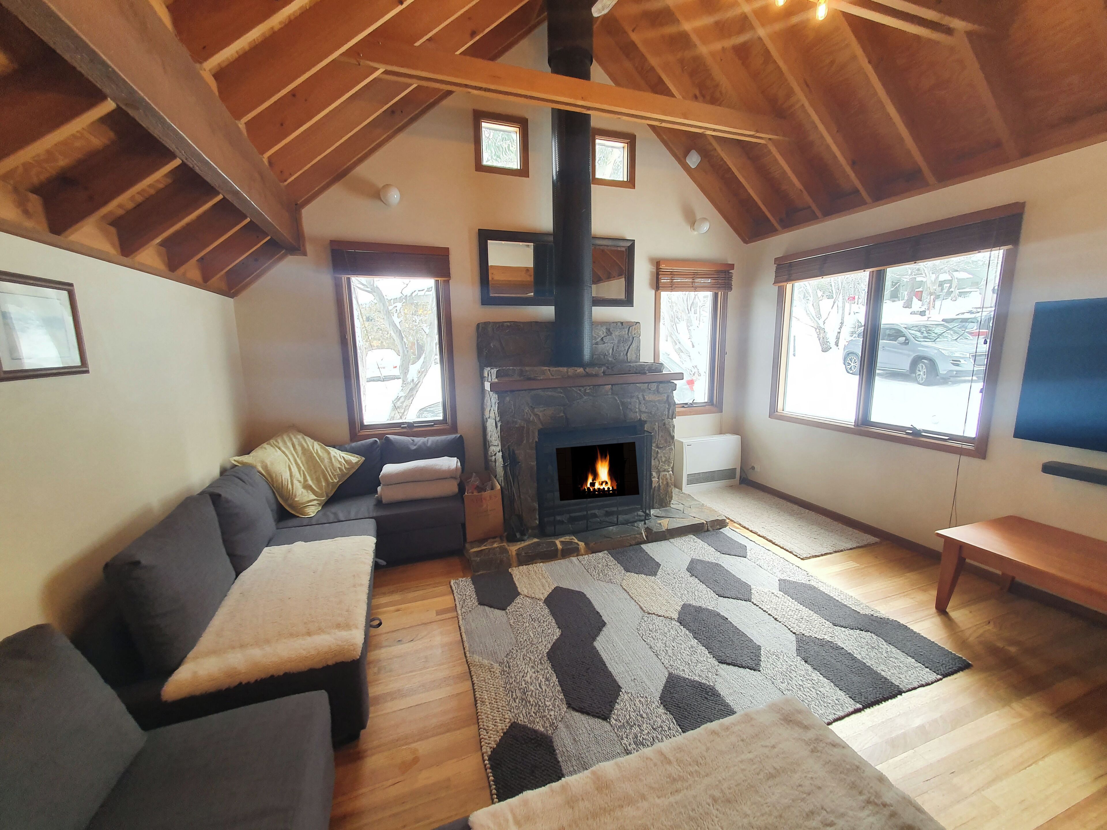 A cozy living room with a stone fireplace, wooden beams, a sectional sofa, and large windows.