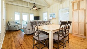 A spacious room with a wooden dining table, four chairs, a ceiling fan, a television, and a large window with blinds. - Laguna Harbor Cottage (Bolivar Peninsula)