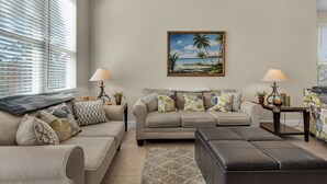 A living room with a beige sofa, a black ottoman, and a painting of a beach scene on the wall. - Happy Ours (Panama City Beach)