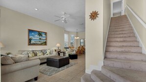 A living room with a sofa, coffee table, and a painting of a beach scene. There is a staircase with carpeted steps leading to an upper floor. - Happy Ours (Panama City Beach)