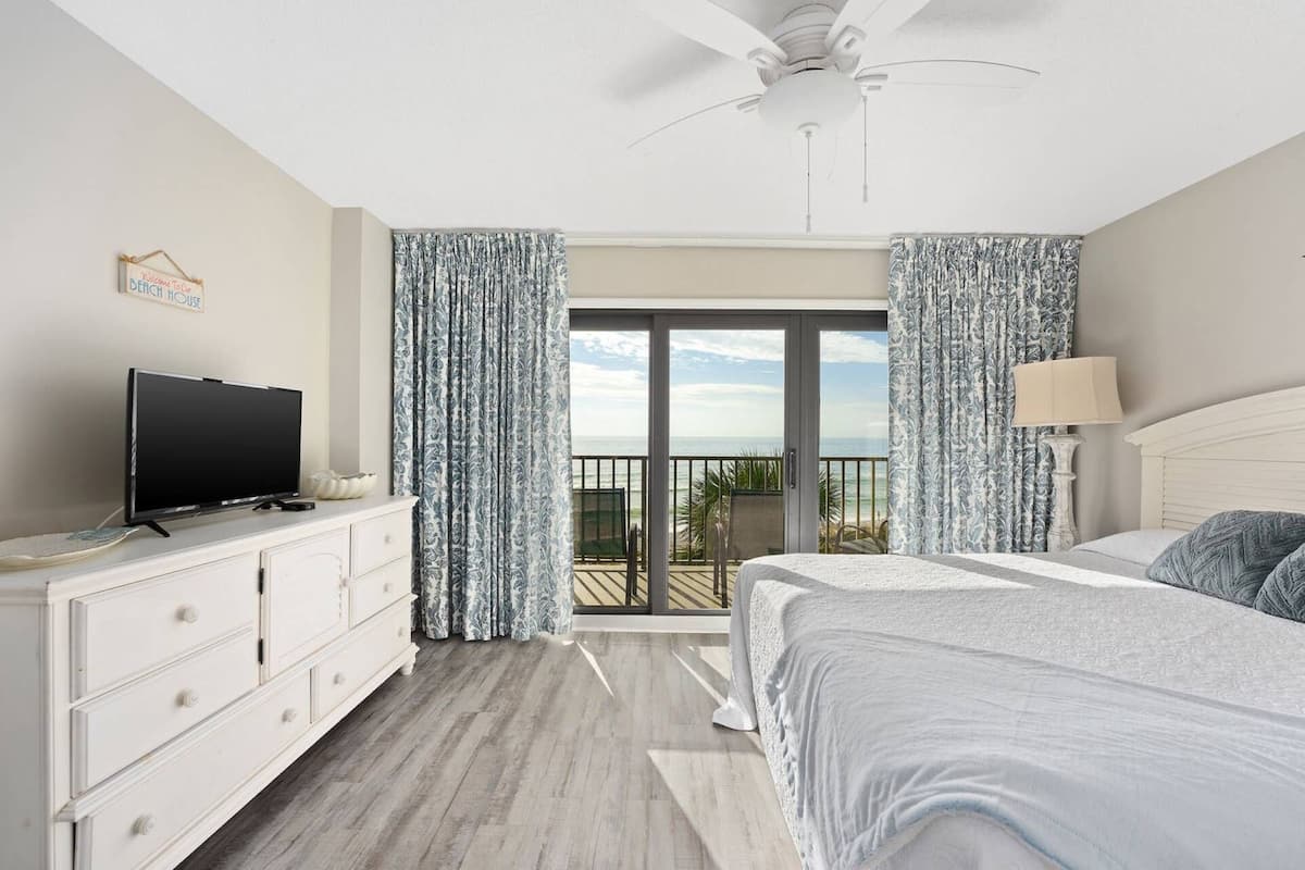 Une chambre à coucher comprenant un lit, une télévision, une commode et un balcon avec des rideaux.