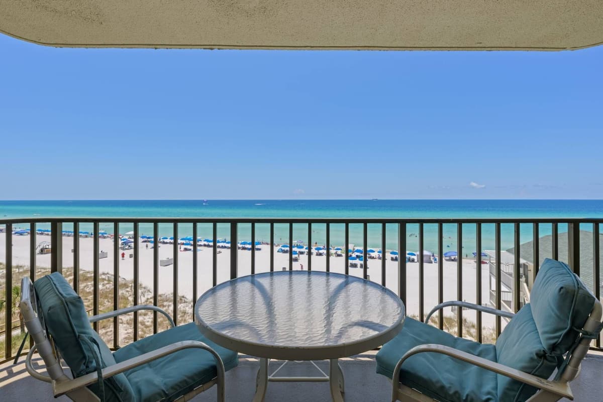 Un balcon avec une table en verre et deux chaises, donnant sur une plage parsemée de parasols et sous un ciel bleu azur.