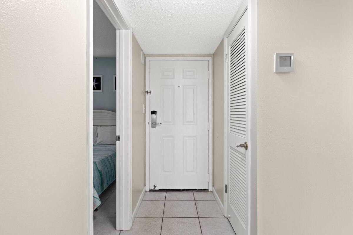 Un couloir étroit avec une porte blanche, un thermostat fixé au mur et un lit dont on aperçoit la couverture à rayures à travers une porte entrouverte.