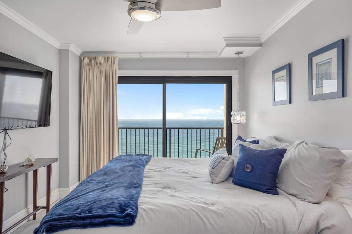 Une chambre d’hôtel équipée d’un lit, d’un bureau, d’une chaise, d’une grande fenêtre donnant sur la mer et d’un ventilateur de plafond.