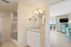 A bathroom with a glass shower enclosure, a sink with a marble countertop, and a large mirror with wall-mounted lights. - Commodore by iTrip Panama City Beach (Panama City Beach)