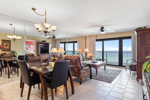 A spacious living room with a dining area, a ceiling fan, and a large window with a view of the ocean. - Commodore by iTrip Panama City Beach (Panama City Beach)