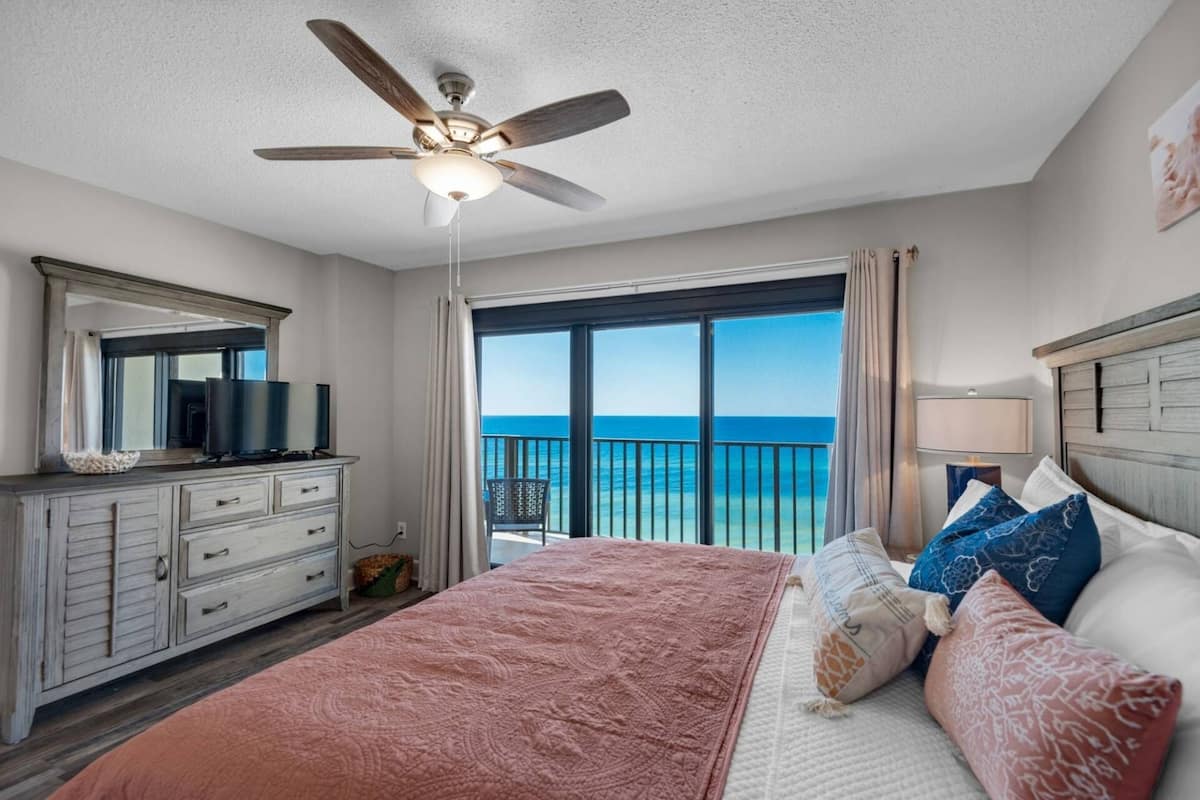 Une chambre avec un lit, un ventilateur de plafond, une télévision, une commode et un balcon avec vue sur l’océan.