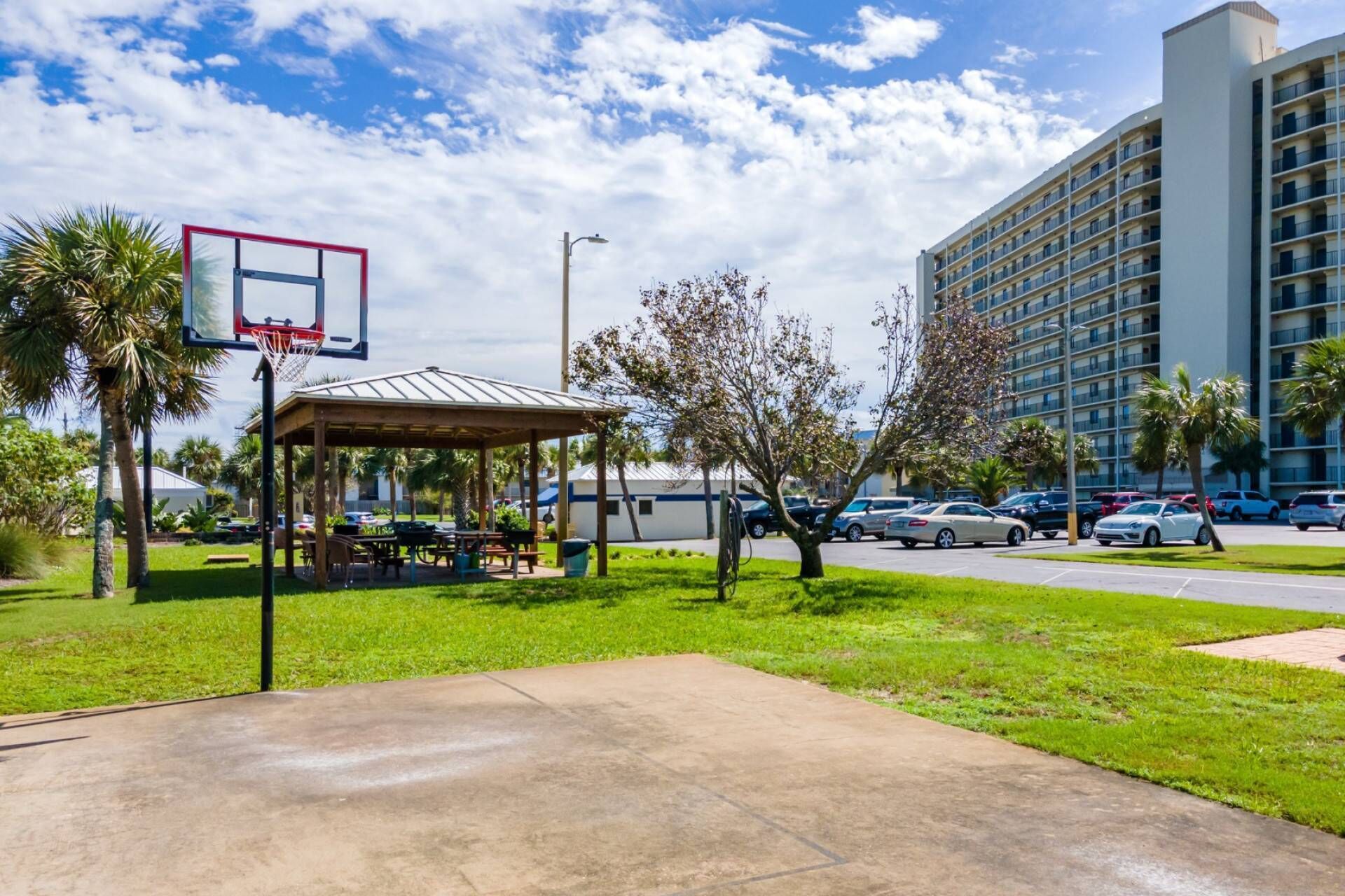 Una cancha de baloncesto al aire libre con un pabellón, palmeras y un estacionamiento con coches.