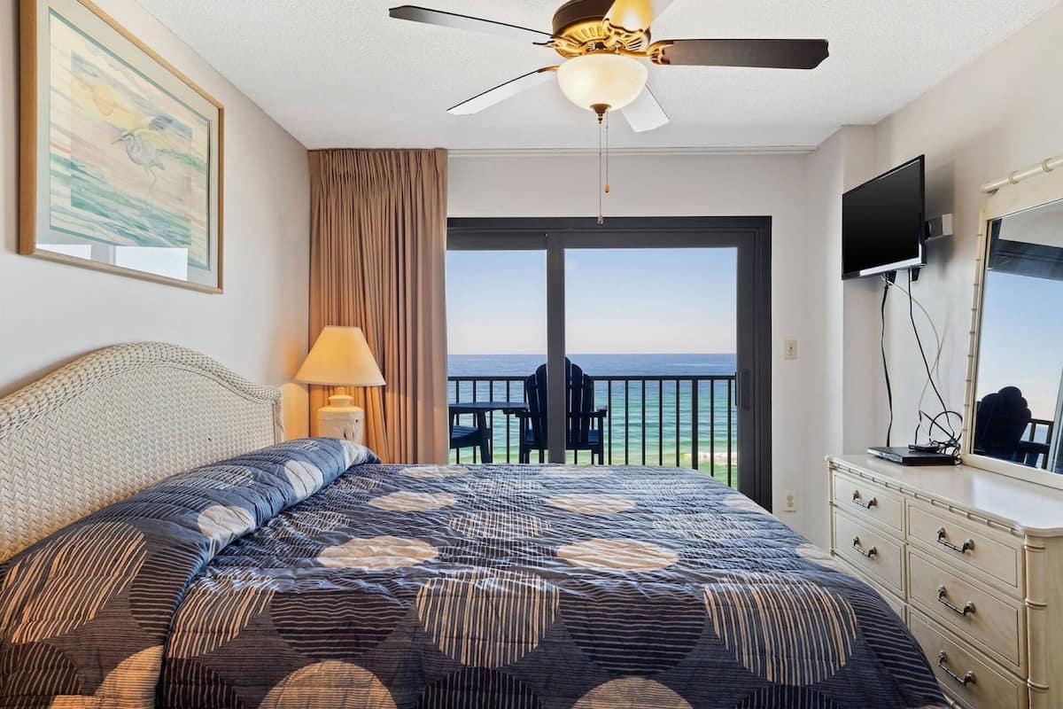 Une chambre d’hôtel avec un grand lit, un ventilateur de plafond, une télévision, une commode et un balcon offrant une vue sur l’océan.