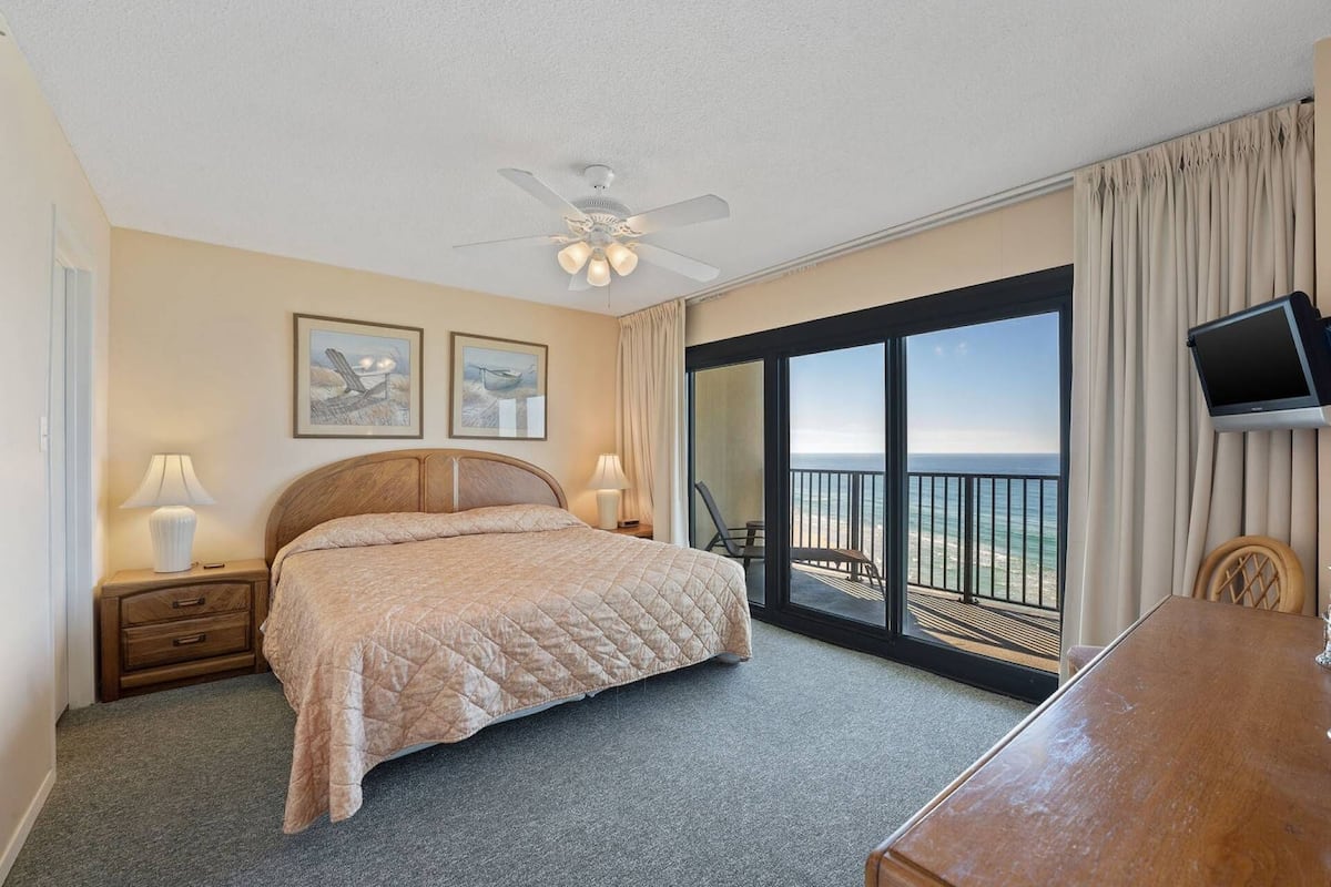 Une chambre d’hôtel équipée d’un lit, d’un bureau, d’une chaise, d’une télévision, d’un ventilateur de plafond et d’un balcon avec vue sur l’océan.