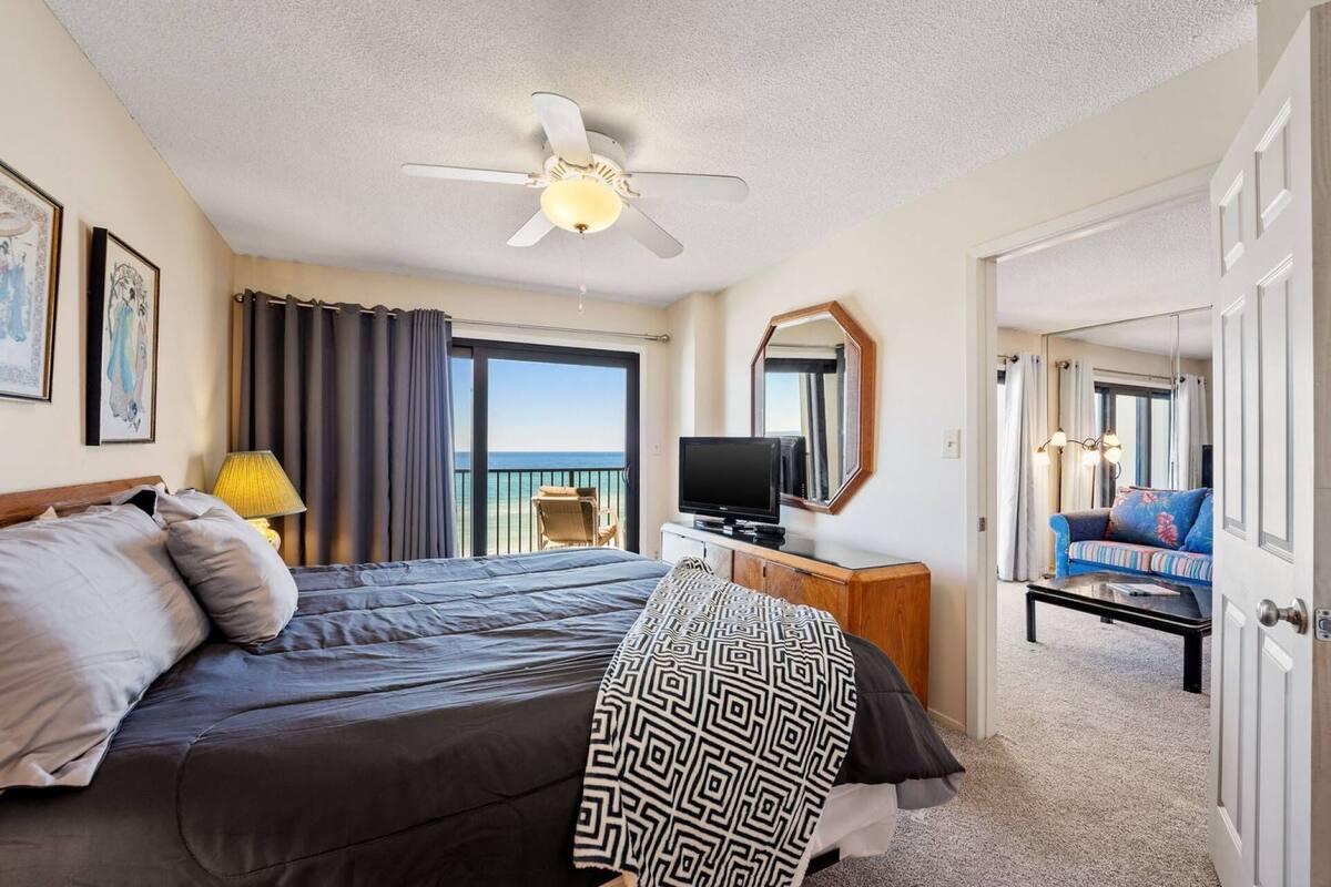 Une chambre d’hôtel comprenant un lit, une télévision, un ventilateur de plafond, un balcon avec vue sur la mer et un coin salon avec un canapé et des fauteuils.