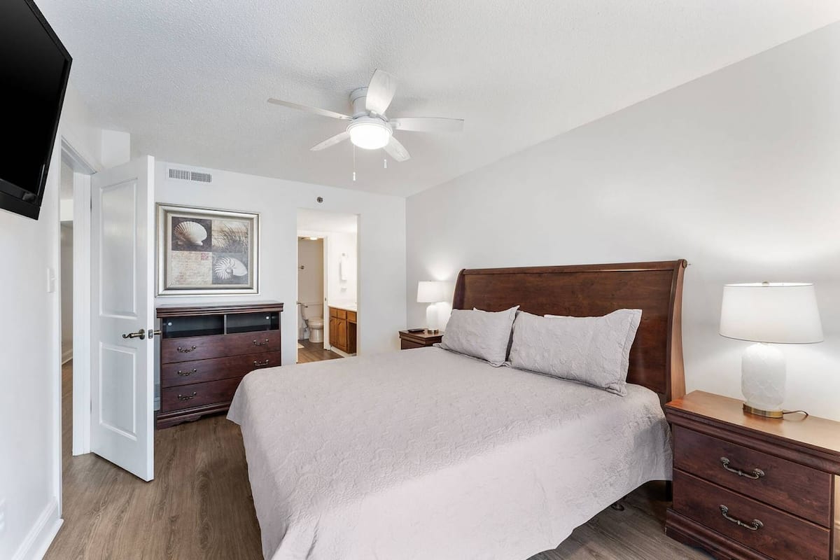 Une chambre à coucher avec un lit, une tête de lit en bois, des lampes de chevet, un ventilateur de plafond, une commode et un tableau encadré au mur.