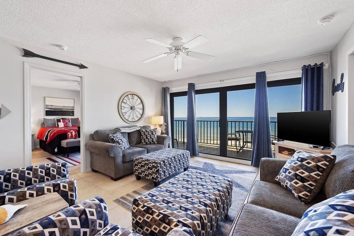 Un vaste salon doté d’une grande fenêtre offrant une vue sur la mer, d’un ventilateur de plafond et d’un téléviseur à écran plat posé sur un meuble en bois.
