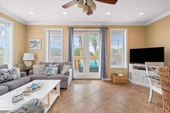 A living room with a sofa, coffee table, TV, and a balcony with a view. - All Decked Out - 140 Southfield (Panama City Beach)