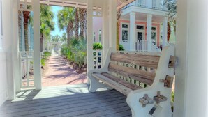 A covered porch with a wooden bench, a white railing, and a view of a pathway leading to a building.