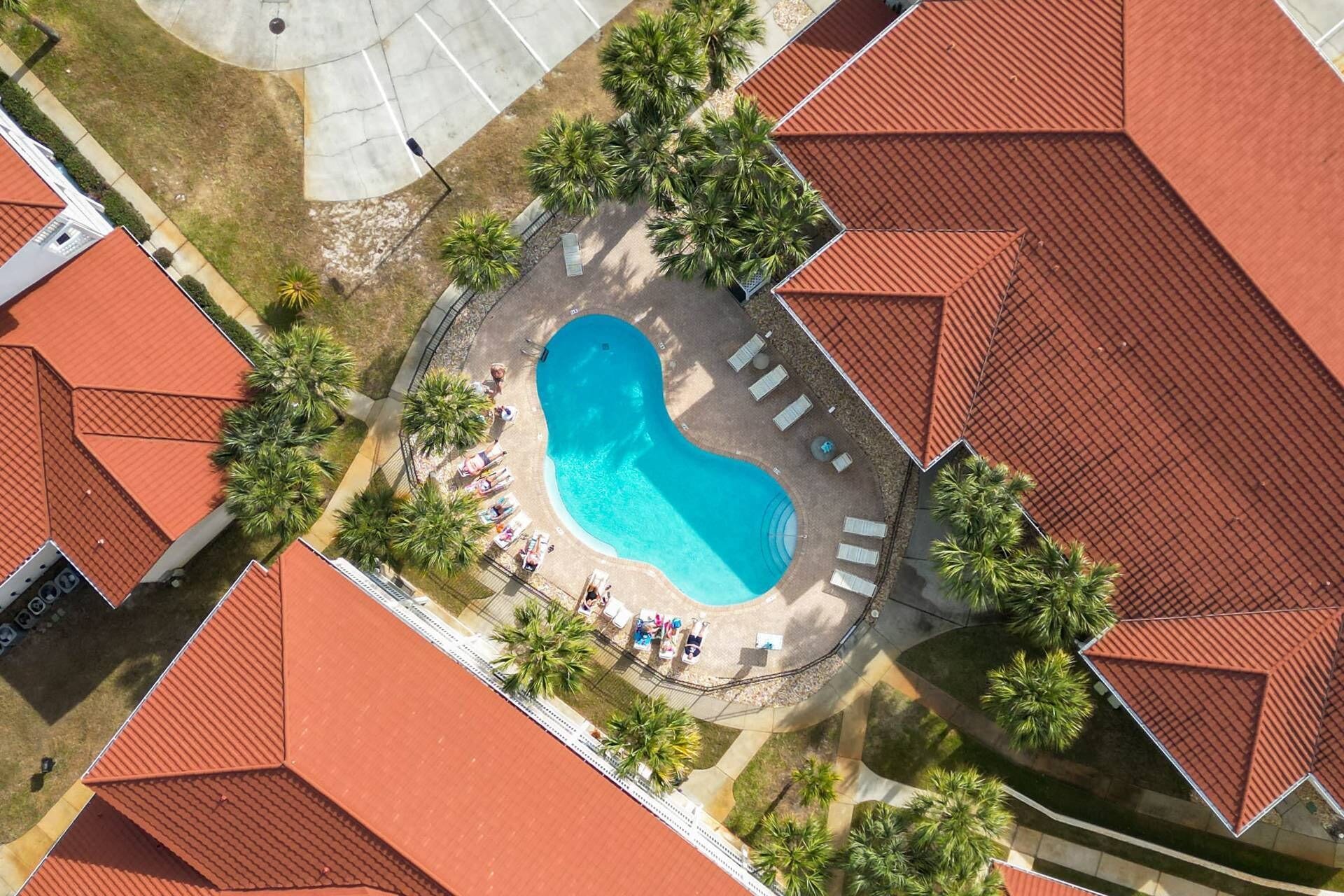 An aerial view of a resort with a central swimming pool surrounded by lounge chairs and palm trees.
