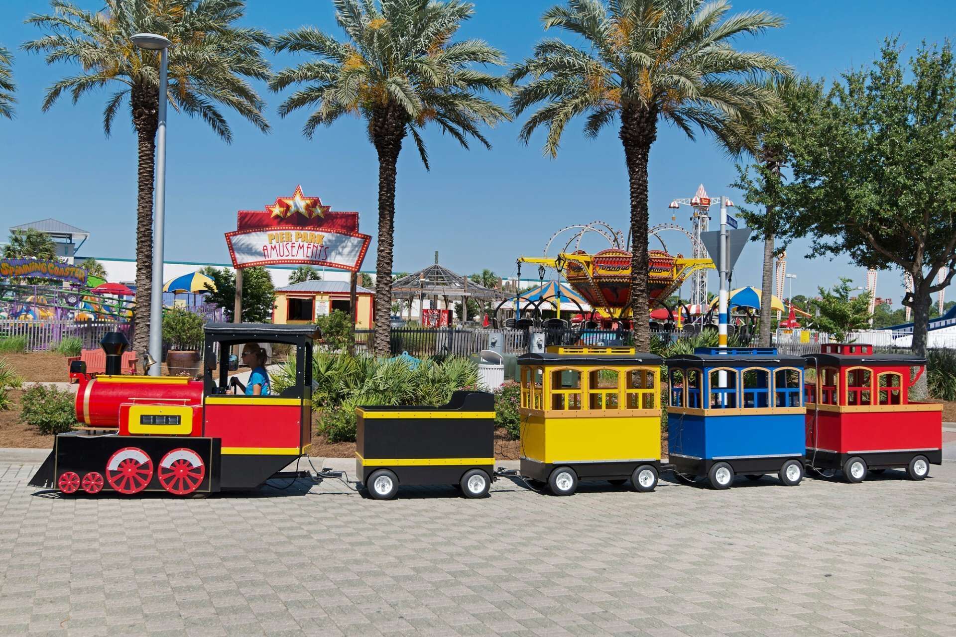 A miniature train ride with colorful carriages, a Ferris wheel, and palm trees in the background.
