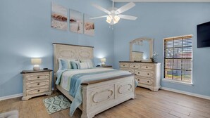 Un dormitorio con cama, cómoda, mesitas de noche, ventilador de techo, obras de arte en la pared y una ventana con persianas.