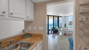 A kitchen with a stainless steel sink, a 'Happy Hour' sign, and a view of a balcony with outdoor seating. - Sunbird Beach Resort 1211 E (Panama City Beach)
