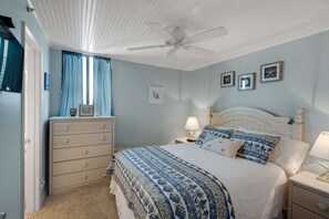 Un dormitorio con cama, mesitas de noche, cómoda, ventilador de techo y ventana con cortinas.