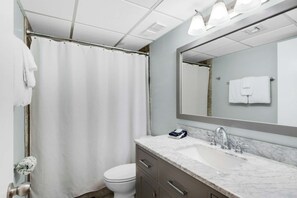 A modern bathroom with a shower curtain, a toilet, a sink with a marble countertop, and a large mirror. - Watercrest by iTrip Panama City Beach (Panama City Beach)