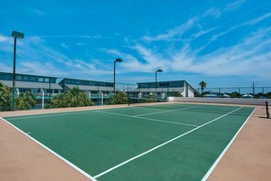 A tennis court with a green surface, surrounded by a fence, and adjacent to a building with multiple windows and balconies. - Watercrest by iTrip Panama City Beach (Panama City Beach)