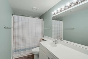 A bathroom with a shower curtain, a toilet, a sink, and a mirror with lights. - Shores of Panama by iTrip Panama City Beach (Panama City Beach)